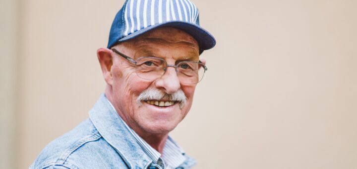 Image shows an older man with a gray mustache and brown-rimmed glasses. He's wearing a blue baseball styple cap with blue and white stripes in the front, and a light blue jeans-type jacket. His expression is pleasant.