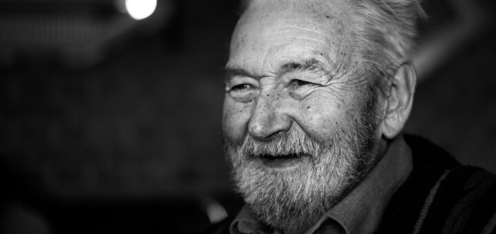 Black and white image shows a night-time background with some lights. Foreground shows a closeup of an older, gray-haired many with a smile and crinkled eyes. He's wearing dark clothes.