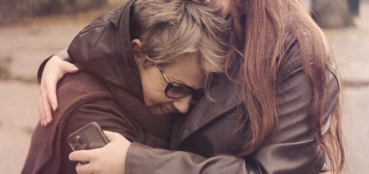 A younger woman with long brown hair smiles as she hugs an older gray-haired woman, possibly her grandmother. Both women are weathering shards of brown.,