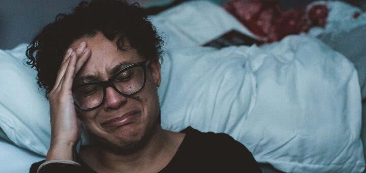 Middle-aged woman lies back abainst white covered pillows. The's got short dark hair and large, dark-rimmed glasses and is wearing a dark pull-over sweater. Her hand it held up by her forehead and her expression is one of misery.