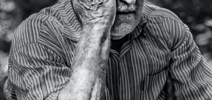 Black and white image shows a very old man in a gray-toned sweater. He's seated and holding his hand up to his face, palm in. His expression shows confusion and fear.