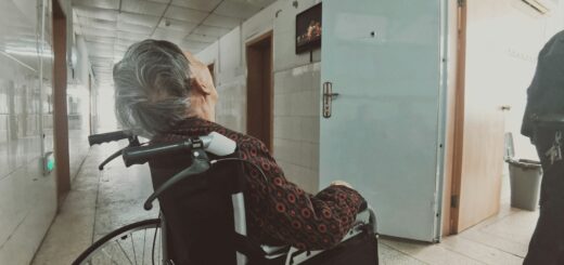 A frail looking older adults in a wheelchair, photographed from the side. Their gray hair is short, and more visible than the face which is turned in the other direction. They are wearing a dark brown sweater.
