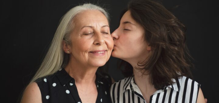Image background is black. On the left is an older woman with long gray hair wearing a dark sleeveless dress with white polka-dots. She's smiling as a younger dark-haired woman wearing a black and white vertically striped blouse kisses her cheek.