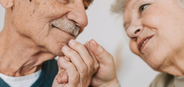The image shows and older coupld against a white background. They are gazing at each other lovingly with hands, held up near their faces, touching as the man clasps the woman's hand. The man is bald with a gray mustache and is wearing a white undershirt topped by a dark blue V-neck. The woman is wearing a beige blouse or sweater.