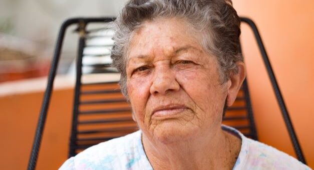 An older woman with salt and pepper hair and brown skin sits in what appears to be a balcony chair that's black wrought iron. She's wearhing a white top with pink and green muted flowers and her expression is unhappy as though complaining.