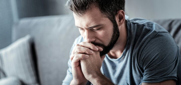 Image shows a younger man with dark hair and a trimmed beard. His eldows are resting on a table, with his hands folded in fron of his mouth. He's leaning into his hands with his eyes closed, as if thinking hard or praying.