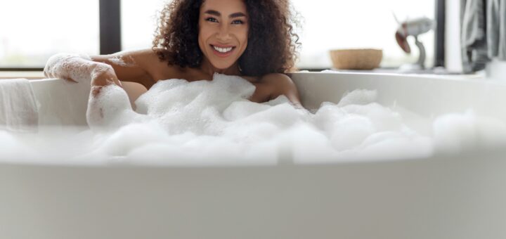A woman with curly dark hair and a smile is luxuriating in a large white tub filled with bubbles.