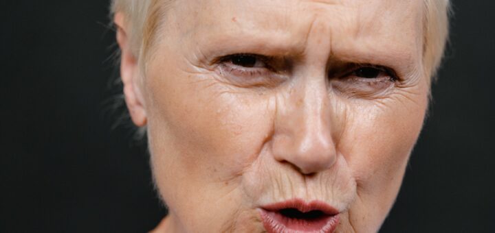 The image background is black. In the foreground, we see a closeup of an older woman's face. She has closely cropped white hair and is wearing a white tee shirt. Her expression shows pursed lips, narrowed eyes, and an angry expression.