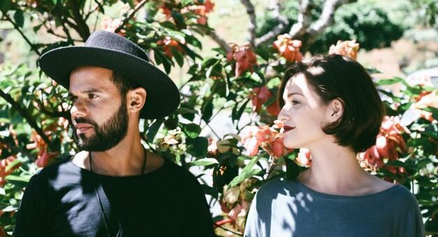 This outdoor image shows a man and a woman in the sun with flower bushes behind. The man wears a black hat and dark tee shirt. He has a dark brown beard. The woman has short drak hair and is wearing a gray/blue shirt. The are both looking to the side at something beyond the camera.