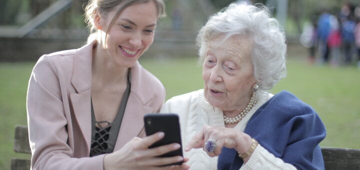 An outside setting shows grass and trees with a large building in the background. A younger woman in a pink sweater with dark blonde hair clipped back is showing an older soman something gon her cell phone. The older woman has gray hair and is wearing a white sweater with a blue shawl over one shoulder.