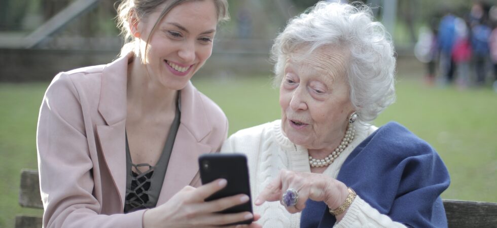An outside setting shows grass and trees with a large building in the background. A younger woman in a pink sweater with dark blonde hair clipped back is showing an older soman something gon her cell phone. The older woman has gray hair and is wearing a white sweater with a blue shawl over one shoulder.
