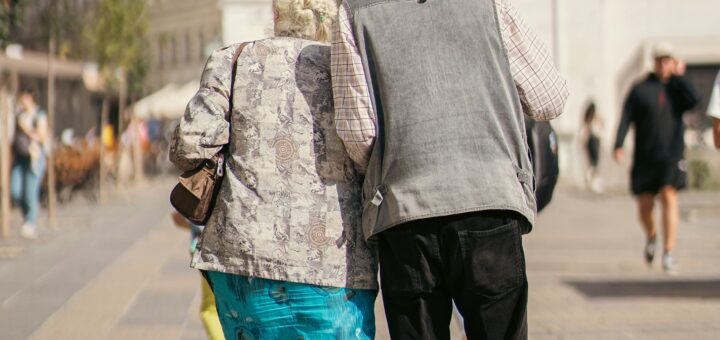 The outdoor image shows a large, white city building in the background. In the foreground, an old couple is shown from the back, walking away from the camera. They are helping each other along as they walk. The man has gray hair, is wearing a gray sweater and black pants, and is slightly bow-legged. The woman is wearing a beige print jacket and a teal and blue print skirt. Her hair is also gray and she has a purse slung over her shoulder.