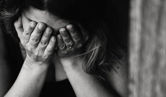 This black and white image shows a close view of a woman with downturned head, hands covering her face in grief. She has dark hair and Hispanic looks.
