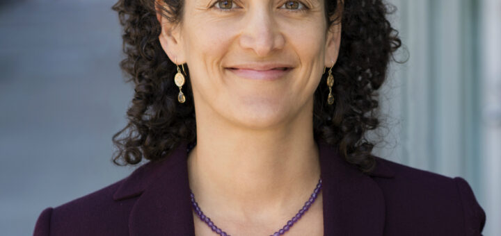 A picture of Dr. Leslie Kernisan. Dr. Kernisan has curly dark hair and is wearing a black suit with a purple bead necklace and small drop earrings. She's smiling in a professional portrait.