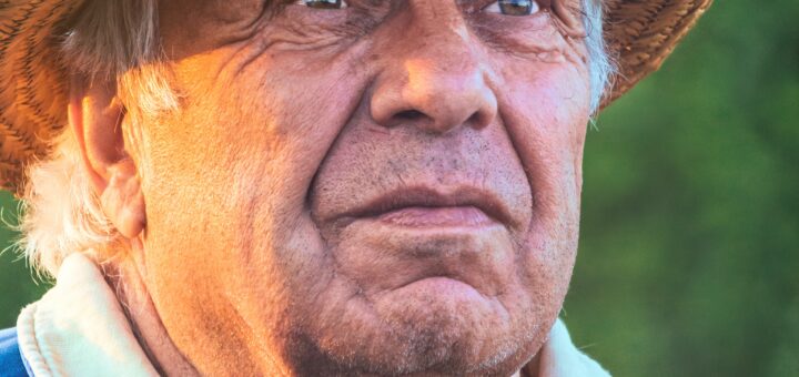 The image shows an older man, maybe Hispanic, wearing a straw hat and blue shirt with a white collar. His expression is contemplative and rather sad as he looks off into the distance.