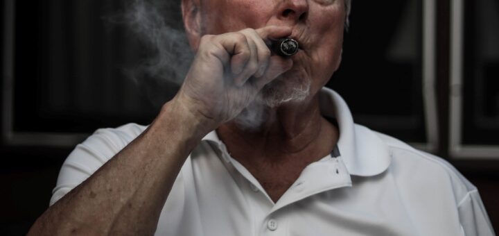 This indoor image shows a confident older man with gray hair smoking a cigar. We see the cigar smoke as he puffs. He's wearing a short-sleeved golf shirt and seems thoughtful but powerful.