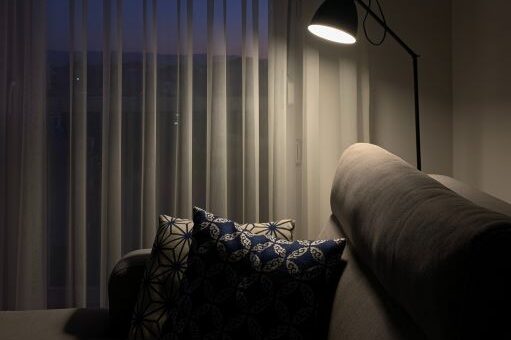 This interior image shows white slatted curtains, a task lamp and a comfortable chair. It denotes comfort while also feeling institutional.