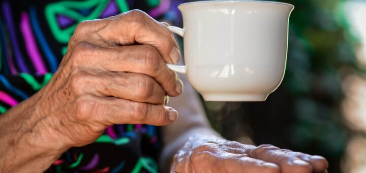 The foreground of the image shows older female hands, one holding a white cup for tea or coffee. The person is wearing a print dress with a black background and lots of color in Green and red, much like Christmas. The blurred background could be a Christmas tree.