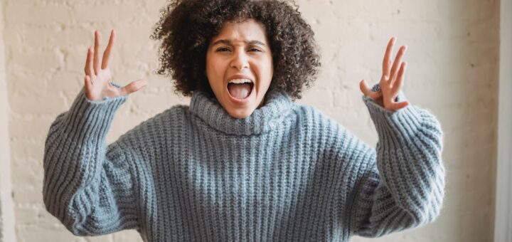 The background is an indoor wall in muted cream. Foreground is a frustrated woman with an Afro that is parted on the side. She has brown skin and is wearing a light tourquoise ribbed turtleneck sweater with long sleeves. Her elbows are bent and hands are held up in the air in frustration or anger.