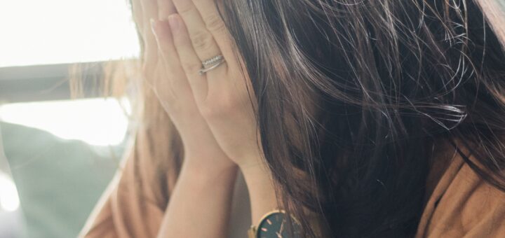 This indoor image shows a closeup of a woman with long brown hair holding her hands over her face. She's wearing red nail polish, wedding rings, and a black modern watch along with a rust colored sweater or wrap visible on her shoulders. She appears to be crying or at least frustrated.