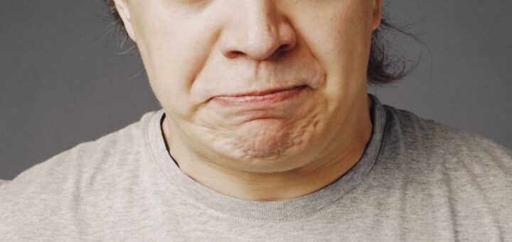 The image shows a closeup of a man with brown hair wearing a beige tee shirt. Heis face wears a bemused expression with raised eyebrows, blue eyes wide open, and mouth showing puzzlement.