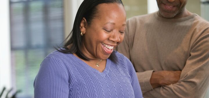 a middle aged African American couple is show happily working in the kitchen. The background shows a home interior. The woman has long, straight black hair and is wearing a lavender sweater. She appears to be peeling a vegetable. The Man is on her left. He has dark hair with a receding hairline. He is wearing a beige sweater and has his arms crossed. Both people are smiling happily.