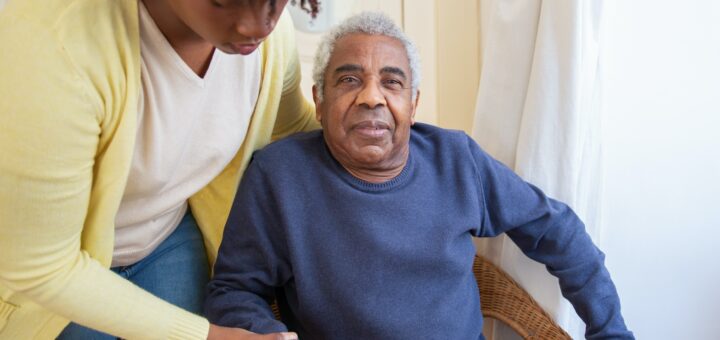 This indoor image shows closed white draperies in the background. ONe the left we see a younger black woman who wears a yellow cardigan over a cream top and jeans. Seh's bending over as she helps an older black man sit in a chair. He has gray hair and is wearing a medium blue pullover.