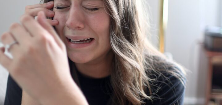 The indoor background shows a blurred gold cross. In the foreground, a woman with long brown hair is crying. Her arms are bent upward with her hands held as if in prayer but also wiping away tears.