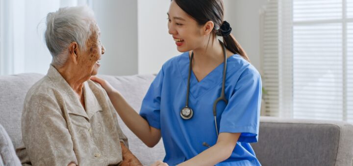 The indoor setting shows two women sitting on a beige couch with white alls in the background. One the left is an older woman with her head turned. She has short gray hair and is wearing a cream colored blouse. By her side is a young woman with long brown hair in a low ponytail. She is wearing medium blue scrubs and has stethoscope hanging around her neck.