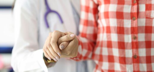 The closeup image only shows the torsos of the people. ON the left is a medical person with a purpose sthoscope around their neck. The person is holding their arm bent and hand out to support a women show is weraing a red and white checked, long-sleeved blouose. She has her hand clasped over the supporting person's wrist.