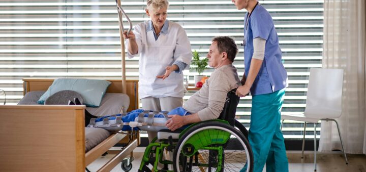 The image shows a hospital room with white blinds closed in the background. Int he foreground, a tech is olding an IV line up as another tech wheels a patient with a cast from let to hip back to their bed. The first tech wears a white coat, then second one wear blue scrubs. The man in the chair is wearing a beige gown or robe.