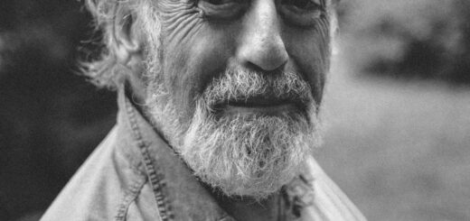 This black and white image has a slightly blurred outdoor background. In the foreground, we see an older man looking directly at the camera. He is balding but has wavy hair sticking out on the sides above his ears and wears a beard. Hair and beard are white. HIs skin is wrinkled and he wears a bemused expression. His shirt is a non-descript collared variety.