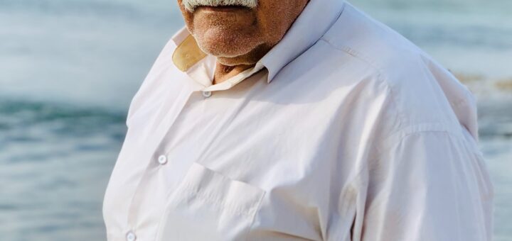 This outdoor image shows a lake in the background with foliage on the other side. The foreground shows an older man, maybe Middle Eastern decent, with white hair and a white mustache. He wears sunglasses and an off-white long-sleeved shirt. His expression is one of displeasure or stubbornness.