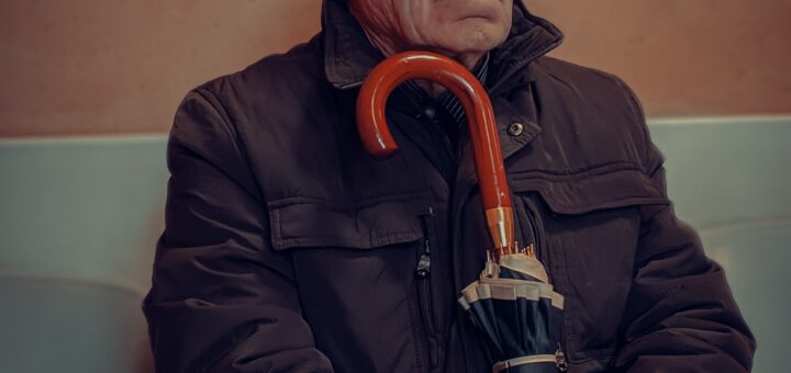 Older man with gray hair and glasses sits on a bench outside. He's wearing a brown coat and holding a folded umbrella with its red handle close to his chest. His expression is stubborn with a set frown.