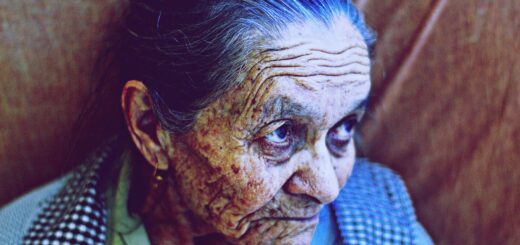 An indoor image of a very old woman with dark, gray-steaked hair pulled back. She is exceptionally wrinkled. She looks to her side with her eyes looking up at someone. Dark shadows underscore her eyes. She wears a black and white checked apron type top over a green blouse.