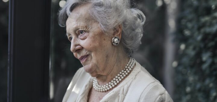 This image shows a blurred indoor background in black and gray. The foreground shows an older woman dressed up for an event. She las curled gray hair and is wear a double-stranded pearl necklace with a light yellow, bulky knit jacket. Her expression shows a soft smilt as she seems to be looking at someone off camera.
