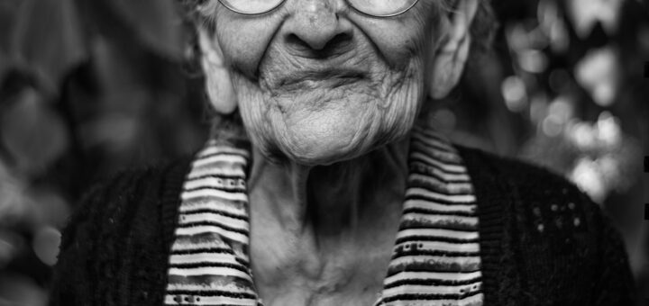 This black and white images shows blurred foliage in the background. In the foreground, a very old woman looks at the camera. She has curly gray hair and round glasses and wears a dark sweater over a striped shirt. Her exopression is one of stubbornness.