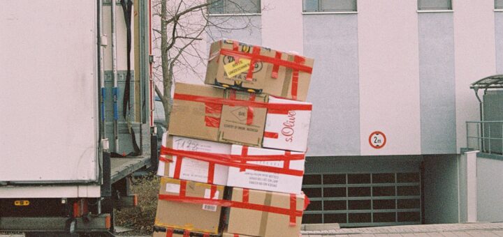 This outdoor image shows a large, two-story white building in the background. The foreground shows the side view of the back of a white moving van. The gate is open and there is a pile of brown cardboard boxes sealed with red tape sitting on the lowered gate.