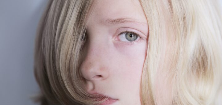 This closeup image shows a young woman with light blonde hair. fair skin, and blue eyes. Her hair is cut into a short bob with long front hair over one eye. She's looking to the side so what we see of her face if sad with a slightly reddened nose.
