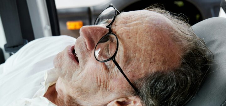 Image shows a closeup of an older man's face. He's lying back on a pillow and appears to be talking with someone. He has salt-and-pepper hair and a receding hairline and wears dark-rimmed glasses.