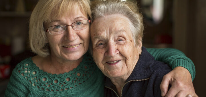 This indoor image shows a blurred background with an older mother-daughter in the foreground. The daughter is one the left. She has a blonde bob, wire rimmed glasses, and wears a green sweater. The mother is on the right. She has steel gray hair drawn back and is wearing a dark, collared top. The younger woman has her arm around the older woman's shoulders and they both wear soft smiiles.
