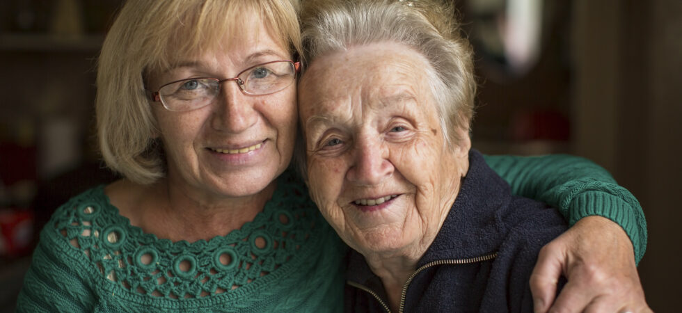 This indoor image shows a blurred background with an older mother-daughter in the foreground. The daughter is one the left. She has a blonde bob, wire rimmed glasses, and wears a green sweater. The mother is on the right. She has steel gray hair drawn back and is wearing a dark, collared top. The younger woman has her arm around the older woman's shoulders and they both wear soft smiiles.
