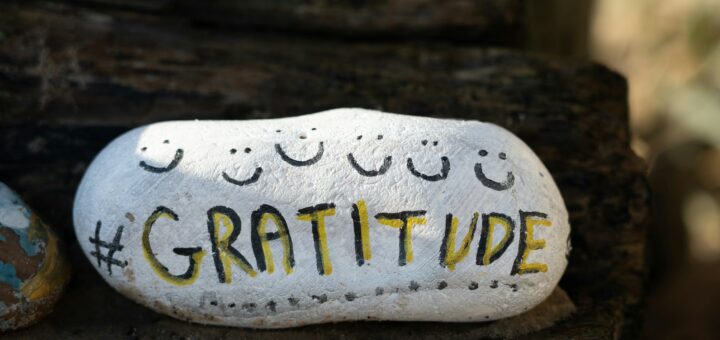 The image shows a dark wood background like a rought table. Stting against this background is a white oblong rock with rounded corners and little smiling faces drawn on in black. Int the center, someone printed in block letters: GRATITUDE.