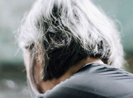 This image has a cloudy, rainy background. We see a woman with bobbed gray hair from the back. Her gray-clad shoulders are bent forward in grief..