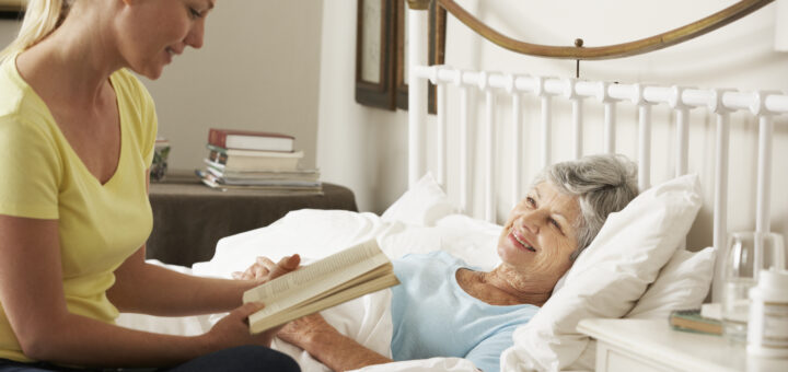 This indoor image shows an older woman lying in a bed with a white frame, her head propped on pillows. She wears a blue nightgown and is smiling up at a younger woman who is reading to her. The younger woman is barely visible, but her profile shows brown hair pulled back and a yellow short-sleeved shirt. She, too, has a pleasant smile on her face as she reads.