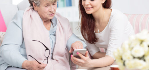 This ndoor image shows a white door with a window blurred in the background. In the foreground, two women site together looking at a smart phone. The older woman has short graying hair, and is wearing a pink wrap around her neck over a blue shirt and jeans. she's holding her glasses in one hand as she points to something on the phone scree that is being held in front of her by a younger woman. The younger woman is on ghe right. She has long grown hair and is wearing a white summery sweater.