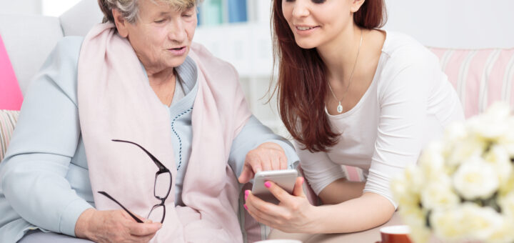 This ndoor image shows a white door with a window blurred in the background. In the foreground, two women site together looking at a smart phone. The older woman has short graying hair, and is wearing a pink wrap around her neck over a blue shirt and jeans. she's holding her glasses in one hand as she points to something on the phone scree that is being held in front of her by a younger woman. The younger woman is on ghe right. She has long grown hair and is wearing a white summery sweater.