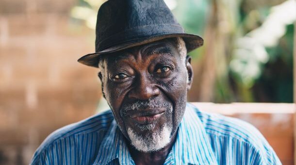 The background is blurred trees. IN the foreground, we see a dapper older black man wearing a black fedora with a blue and white striped collared shirt. He has a bray goatee with white sideburns and a bemused expression.