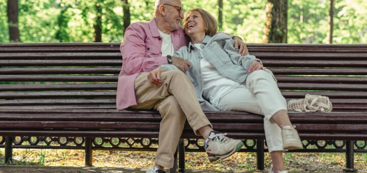 An older couple sit close on a park bench with trees in the background. The man wears a pink sport coat, khakis, and sneakers. He's bald, wears glasses, and has his arm around the woman who looks up into his face with a smile. The woman has graying brown hair and wars a light green cardigan and cream pants with sneakers.