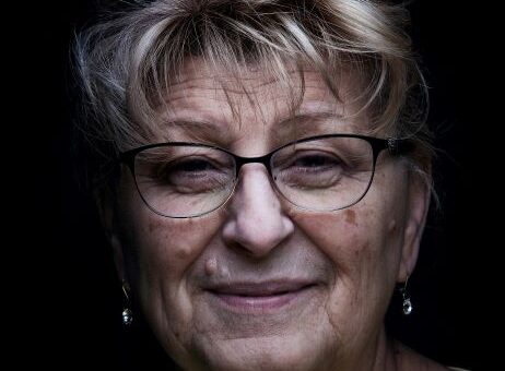 The background is black. In the foreground we see an older woman's face close up. She has salt and pepper hair that's been curled in front with bangs, and she wears dark rimmed glasses. Her expression is one with a soft smile, maybe slightly confused.